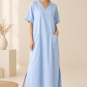 May include: A light blue maxi dress featuring a V-neckline, short sleeves, two large front patch pockets, and side slits at the hem. The garment is worn with a light-colored head covering and simple slide sandals.