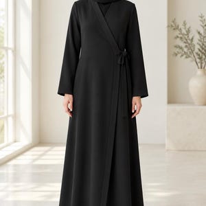 May include: A solid black wrap-style abaya featuring long sleeves, a V-neckline, and a side tie closure at the waist. The garment is floor-length and is paired with a matching black hijab that covers the head and neck area completely.