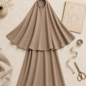 Może przedstawiać: Beżowy wykrój krawiecki PDF Jilbab Noor w rozmiarach od S do XL. Odzież składa się z peleryny z kapturem i pasującej spódnicy. Obok znajduje się miarka krawiecka, nożyczki oraz papierowa ilustracja przedstawiająca projekt wzoru.