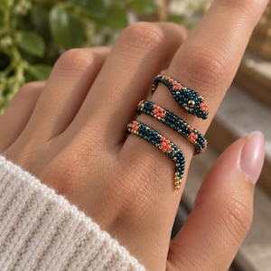 May include: A coiled snake ring crafted from small beads in deep teal and coral pink. The snake head features a single gold-toned bead for an eye, with the body wrapping around the finger in a spiral design, showcasing intricate beadwork.