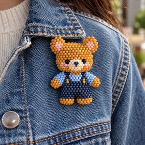 May include: A beaded bear brooch featuring a brown bear with pink inner ears and cheeks, wearing light blue sleeves and dark blue overalls. The bear is constructed from small, uniform beads and is attached to a blue denim jacket.