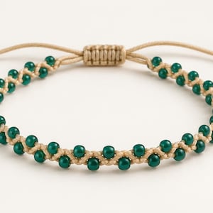 May include: A bracelet featuring a beige cord woven with small, round, dark green beads. The design includes an adjustable sliding knot closure with two loose cord ends, each finished with a single dark green bead at the tip.