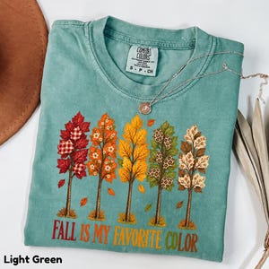 Może przedstawiać: Jasnozielony t-shirt z grafiką przedstawiającą pięć stylizowanych drzew z liśćmi o różnych wzorach, w tym w kratę, kwiaty i cętki lamparta. Poniżej drzew znajduje się pomarańczowy napis "FALL IS MY FAVORITE COLOR".