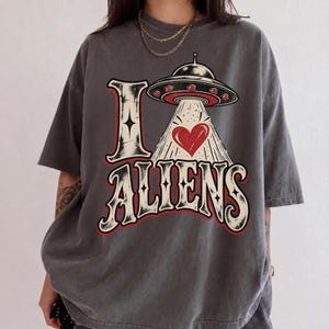 May include: A charcoal grey short-sleeved t-shirt featuring a graphic print that reads I LOVE ALIENS. The design includes a flying saucer beaming down a red heart, with the letter I replaced by a star and the word ALIENS in a bold font.