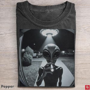 Könnte beinhalten: Ein anthrazitfarbenes T-Shirt mit einem schwarz-weißen Grafikdruck eines Außerirdischen vor einer Türklingelkamera. Der Außerirdische hält eine brennende Zigarette, während ein leuchtendes UFO am Nachthimmel über einer Einfahrt schwebt.
