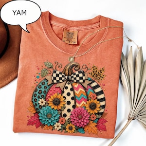 May include: An orange t-shirt with a graphic of a pumpkin decorated with colourful patterns including leopard print, polka dots, and zigzags. The pumpkin is adorned with a black and white chequered bow and various flowers like sunflowers and dahlias.