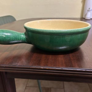 May include: A round ceramic bowl with a single handle extending from the side. The exterior of the bowl features a glossy dark green glaze, while the interior is finished in a smooth, light cream-colored glaze with a subtle speckled texture.