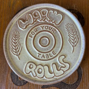 May include: A round, beige ceramic plate with a glossy finish. The text "WARM FOR YOUR TABLE ROLLS" is embossed in a stylized font. Two leaf-like illustrations are positioned on the left and right sides of the central circular design.