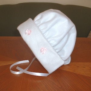 May include: White fleece baby bonnet with two pink satin bows.