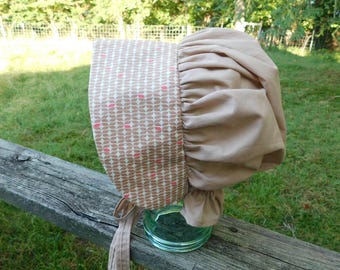 May include: A beige and patterned bonnet with a floral design. The bonnet features a patterned front panel with a repeating pattern of brown shapes and pink accents. The back of the bonnet is a solid beige colour, with a ruffled edge and ties.