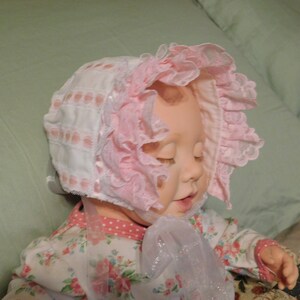 May include: A baby doll wearing a pink and white bonnet with lace trim. The bonnet has a white satin ribbon tied under the chin. The doll is wearing a white and pink floral dress.