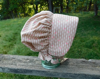 May include: A beige and pink patterned bonnet with a ruffled edge. The bonnet is made of fabric with a repeating leaf-like design in pink and beige. The bonnet is sitting on a glass jar.