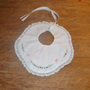 Puede incluir: Babero de bebé de algodón blanco con borde festoneado y flores bordadas. El babero tiene un lazo de cinta de satén blanco.