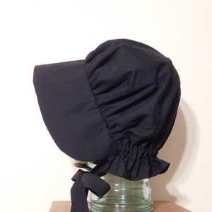 Könnte beinhalten: Eine schwarze Haube mit breiter Krempe und geraffter Krone. Die Haube hat einen gerüschten Rand und eine schwarze Schleife. Der Stoff scheint einfarbig und blickdicht zu sein. Die Haube wird auf einem transparenten Glasobjekt präsentiert.