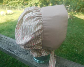 Op de afbeelding: Een beige babykap met een patroon en een effen rand. De stof met patroon heeft een herhalend ontwerp van bruine en rode bladvormen. De kap is vastgebonden met bijpassende linten. De achtergrond is een grasveld.