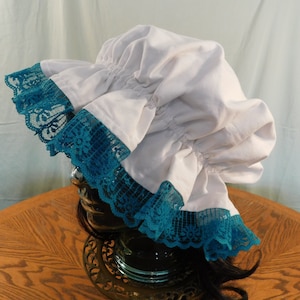 Op de afbeelding: Witte katoenen muts met een geplooide ruche en een teal kantversiering.