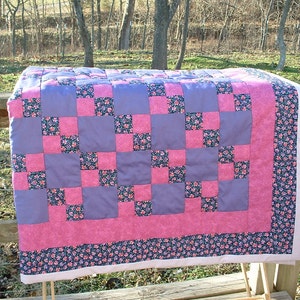 May include: A patchwork quilt with a checkered pattern in shades of pink and purple. The quilt is made of squares of fabric with floral patterns.