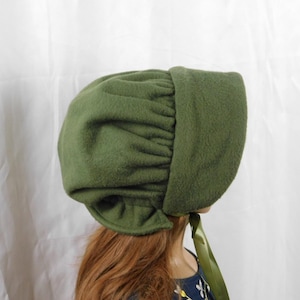 Puede incluir: Gorro verde oliva con una corona redondeada y un ala ancha. El gorro está hecho de una tela suave y texturizada y presenta un diseño fruncido en la corona. Una cinta verde está atada debajo de la barbilla.