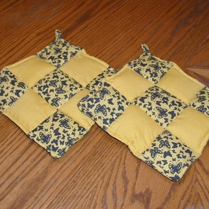 Op de afbeelding: Twee gele en zwarte gequilte ovenwanten met een vlinderpatroon. De ovenwanten hebben een lus om op te hangen.