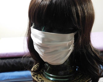May include: White cotton face mask with pleats, worn on a mannequin head with long brown hair.