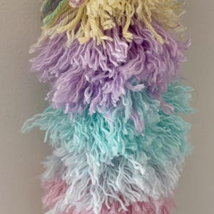 Peut inclure: Un porte-clés pompon décoratif composé de fils de laine multicolores dans des tons pastel de lavande, jaune pâle, bleu clair et rose tendre. La laine est texturée et effilochée, suspendue à un fermoir mousqueton en métal argenté.