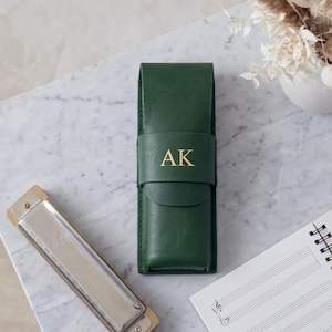 Könnte beinhalten: Ein dunkelgrünes, rechteckiges Etui mit Klappverschluss und den gold geprägten Buchstaben AK auf der Vorderseite. Neben dem Etui liegen eine silber-goldene Mundharmonika und ein Spiralblock mit aufgedruckten Notenlinien auf den weißen Seiten.