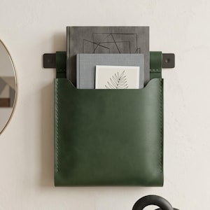 Könnte beinhalten: Ein dunkelgrüner Wand-Organizer aus Leder mit sichtbaren Nähten an den Seiten. Die Tasche enthält ein dunkelgraues Buch, ein hellgraues Buch und eine weiße Karte mit einer schwarzen Strichzeichnung eines Farnblatts an einer Wand.