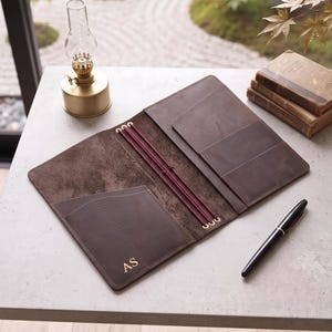 Può includere: Una copertina per diario in pelle marrone scuro aperta con tre elastici verticali color rosso intenso, tasche interne e le iniziali AS impresse in oro nell'angolo in basso a sinistra, accompagnata da una penna stilografica nera.