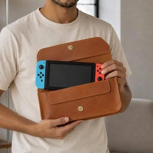May include: A tan leather envelope-style carrying case with a gold snap closure, containing a handheld gaming console with a black screen and detachable blue and red controllers. The case is rectangular with rounded corners and a flap top.
