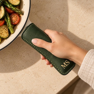 May include: A dark green leather skillet handle cover featuring visible stitching along the edges and the gold embossed initials MS. The cover is attached to the handle of a white enameled cast iron pan containing cooked asparagus and tomatoes.