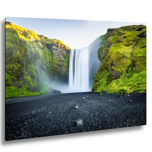 May include: A landscape photograph featuring a tall, vertical waterfall cascading between two steep, moss-covered green cliffs. The base of the waterfall meets a dark, volcanic black sand beach with scattered small rocks under a bright sky.