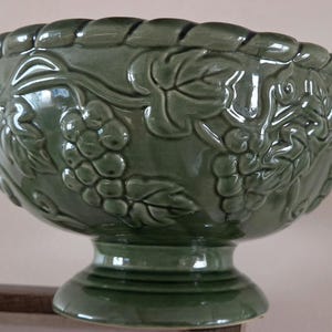 Puede incluir: Un cuenco de cerámica verde salvia sobre pedestal con un patrón en relieve de racimos de uvas y hojas de parra en el exterior. El borde tiene una textura de cuerda decorativa y el cuenco se asienta sobre una base circular firme y brillante.