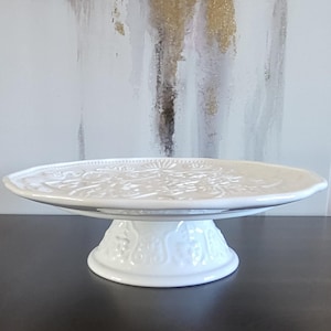 May include: A white ceramic cake stand featuring a raised, embossed floral and scroll pattern on the circular plate and the pedestal base. The stand has a glossy finish and a scalloped edge, providing a raised platform for serving food items.