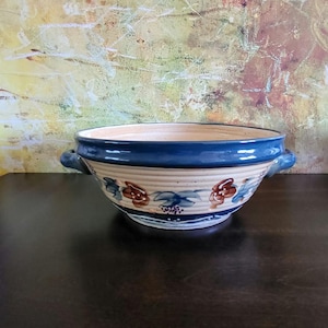 May include: A ceramic bowl with a cream-coloured base and a wide, dark blue rim. The exterior features horizontal ridges and a hand-painted floral pattern in shades of brown, blue, and green. Two small, curved handles are attached to the sides.