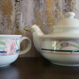 以下が含まれることがあります： ピンクの花、緑の葉、そして濃紺とピンクの水平なストライプが描かれた花柄の白い陶磁器製のティーポットとティーカップのセット。ティーポットには蓋、湾曲した注ぎ口、持ち手が付いており、ティーカップには持ち手が一つ付いています。