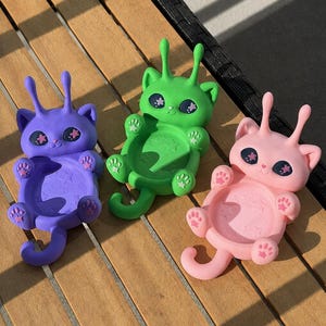 May include: Three cat-shaped phone stands in purple, green, and pink. Each figure features two antennae, large dark eyes with star-shaped pupils, and raised paws. The center of each stand has a circular base embossed with star patterns.
