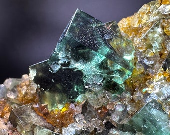 May include: Close-up of a fluorite crystal cluster. The image showcases translucent, cubic fluorite crystals in shades of green and teal, with hints of yellow and brown. The crystals are set against a dark background, highlighting their geometric forms.