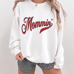 May include: A white crewneck sweatshirt featuring the word Mommin' printed in a bold red script font with a black outline. The garment has long sleeves and a ribbed collar, cuffs, and hem, providing a relaxed fit for casual wear.