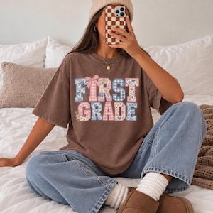 May include: A brown short-sleeve t-shirt featuring the text FIRST GRADE in a block font. The letters have a blue and pink gingham and floral pattern with a small pink bow on the letter F. The shirt is worn with blue jeans and a beige beanie.