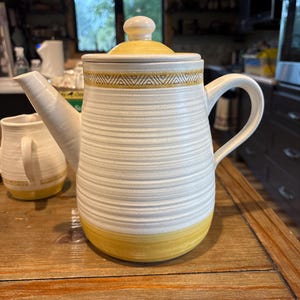 Puede incluir: Una tetera de cerámica con cuerpo color crema, estrías horizontales y base amarilla. La tapa tiene un borde amarillo a juego y un pomo. Un patrón geométrico en amarillo rodea la parte superior de la tetera, que también incluye un asa curva y un pico vertedor cónico.