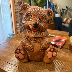 May include: A brown ceramic bear cookie jar featuring a textured fur finish, blue eyes, a black nose, and a small red tongue sticking out. The bear is in a seated position with light pink paw pads on its feet and hands, showcasing a detailed design.