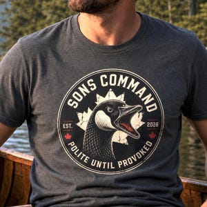 Peut inclure: Un t-shirt gris chiné foncé à col rond avec un motif circulaire représentant une tête de bernache du Canada. Le texte indique SONS COMMAND, EST. 2026 et POLITE UNTIL PROVOKED. Une silhouette de feuille d'érable est visible avec deux petites feuilles rouges.