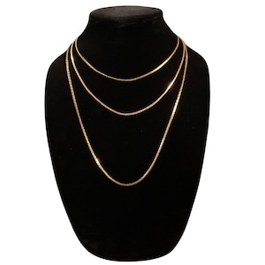 May include: A set of three layered gold-toned chains of varying lengths, each featuring a delicate, flat, reflective link design. The chains are arranged in a cascading formation, creating a tiered appearance with a polished, metallic finish.