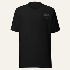 May include: A black short-sleeved t-shirt featuring a small rectangular graphic on the upper left chest. The graphic contains the text "THE LOST NOTEBOOK" in a serif font, enclosed within a thin border with decorative lines above and below.