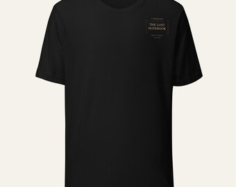 May include: A black short-sleeve t-shirt featuring a small rectangular graphic on the upper left chest. The graphic contains the text "THE LOST NOTEBOOK" in a serif font, enclosed within a thin border with decorative lines above and below.