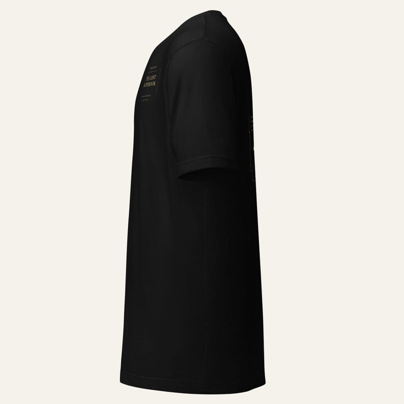 May include: A black short-sleeved t-shirt viewed from the side. The shirt features a small rectangular graphic on the chest area containing the text THE LOST NOTEBOOK in a serif font, enclosed within a thin border with decorative corner accents.