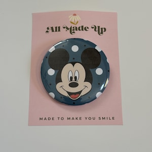 May include: A circular pinback button featuring a graphic of Mickey Mouse on a dark blue background with white polka dots. The button is attached to a light pink display card with the text All Made Up and MADE TO MAKE YOU SMILE.