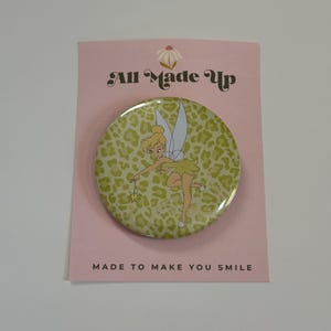 May include: A circular pinback button featuring a cartoon fairy with blonde hair and wings, set against a light green leopard print background. The button is attached to a pink display card with the text All Made Up and MADE TO MAKE YOU SMILE.