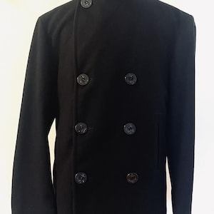 Op de afbeelding: Een effen zwarte dubbelrijige jas met een hoge opstaande kraag en een sluiting aan de voorzijde met zes donkere ronde knopen. De jas heeft lange mouwen en een gestructureerd silhouet, wat zorgt voor een strak en uniform uiterlijk.