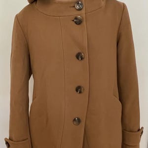 May include: A camel-colored coat featuring a hood with a vibrant magenta interior lining. The coat has a high neckline, a front button closure with four large brown marbled buttons, long sleeves with buttoned cuffs, and two side pockets.